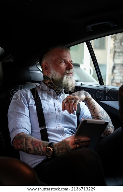 Tattooed old man with tablet sitting inside car\
looking away