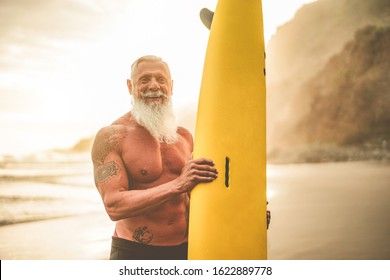 Tattooed Senior Surfer Holding Surf Board On The Beach At Sunset - Happy Old Guy Having Fun Doing Extreme Sport - Joyful Elderly Concept - Focus On His Face