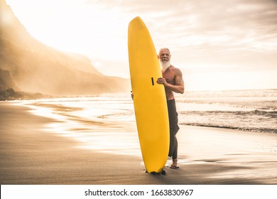 Tattooed Senior Surfer Holding Surf Board On The Beach At Sunset - Happy Old Guy Having Fun Doing Extreme Sport - Joyful Elderly Concept - Focus On His Face