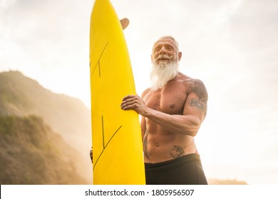 Tattooed Senior Surfer Holding Surf Board On The Beach At Sunset - Happy Old Guy Having Fun Doing Extreme Sport - Joyful Elderly Concept - Focus On His Face