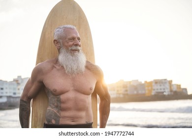 Tattooed Senior Surfer Holding Vintage Surf Board On The Beach At Sunset - Happy Old Guy Having Fun Doing Extreme Sport - Joyful Elderly Lifestyle Concept - Focus On Face