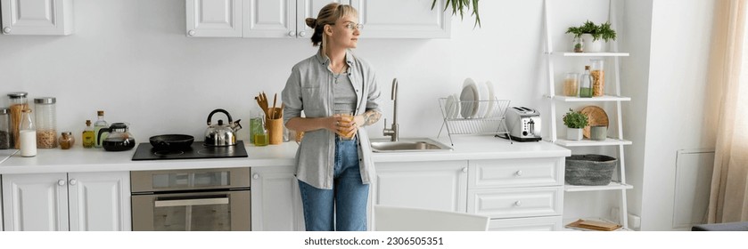 tattooed woman with bangs and eyeglasses holding glass of orange juice and kitchen worktop, kettle,frying pan, coffee pot, clean dishes, toaster, rack with plants, bottle with milk at home, banner - Powered by Shutterstock