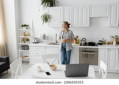 tattooed woman with bangs and eyeglasses holding glass of orange juice and standing near kitchen worktop next to desk with devices, bowl with cornflakes and cup of coffee on saucer at home, freelance - Powered by Shutterstock