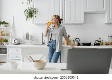 tattooed woman in eyeglasses drinking orange juice and standing near  kitchen worktop next to desk with devices, bowl with cornflakes and cup of coffee with saucer at home, freelance lifestyle - Powered by Shutterstock