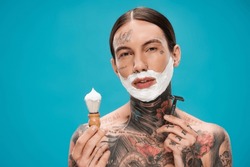 Tattooed Young Man With Shaving Foam On Face Holding Vintage Brush And Safety Razor Isolated On Blue.Translation Of Tattoo:'kiss Passionate And Bite Gently'