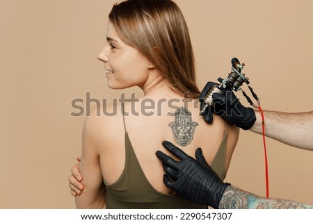 Tattooer master artist hold machine black ink in jar, equipment for making tattoo art on back view smiling happy caucasian woman female body shoulders back isolated on plain beige background studio