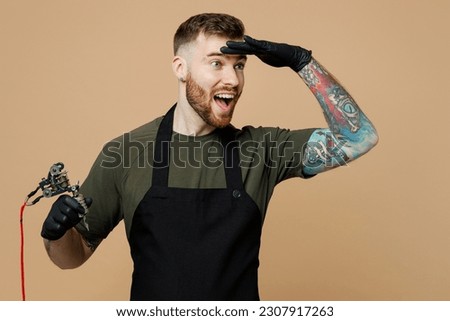 Tattooer master artist man wear green t-shirt apron hold hand at forehead look far away distance, machine black ink in jar, equipment for making tattoo art on body isolated on plain beige background