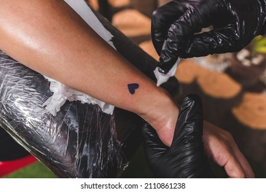 A tattooist cleans the surrounding skin of a fresh heart tattoo on a client's forearm with a tissue. Final procedure. - Shutterstock ID 2110861238