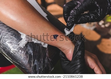 A tattooist cleans the surrounding skin of a fresh heart tattoo on a client's forearm with a tissue. Final procedure.
