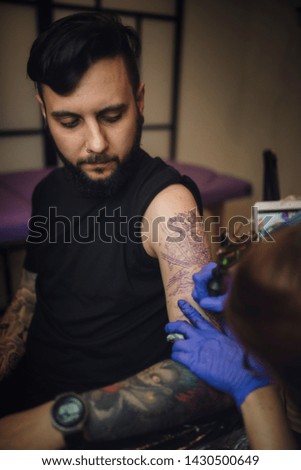 tattooist girl makes a tattoo in her studio to a young guy with a beard