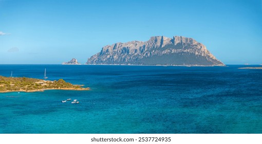 Tavolara Island features dramatic limestone cliffs and turquoise waters. Small boats are anchored near the lush shoreline under a clear blue sky. - Powered by Shutterstock