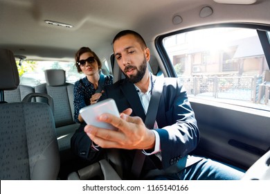 Taxi driver sitting in a car and talking with businesswoman, holding smart phone in hand.
