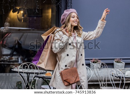 Taxi, I am here! Side view of beautiful blonde woman in beret holding shopping bags in hand and smiling while calling for taxi. Waiting for cab after long shopping day. French woman shopaholic.