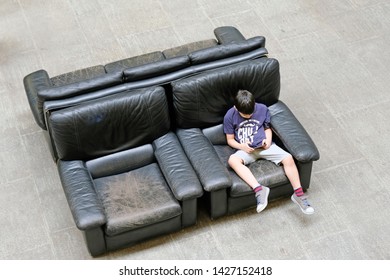 Tbilisi, Georgia - June 7, 2019: A Little Boy Sits Alone On The Couch And Plays The Game On The Phone Top View. Old Black Leather Sofa And A Kid With Phone.