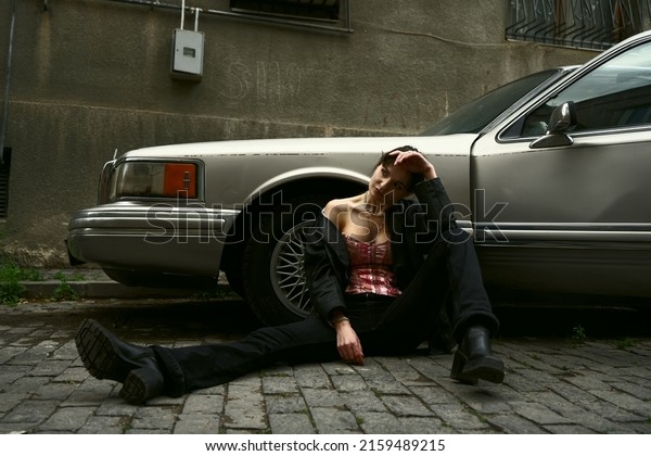 TBILISI,\
GEORGIA - MAY 16, 2022: female model posing  near retro, vintage\
american vehicle - Lincoln Town car. beautiful woman sitting on\
floor. Girl sitting on grey aged paving\
stone