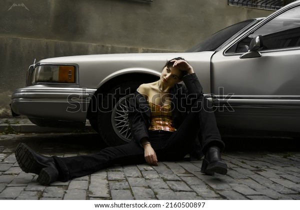 TBILISI,\
GEORGIA - MAY 16, 2022: female model posing  near retro, vintage\
american vehicle - Lincoln Town car. beautiful woman sitting on\
floor. Girl sitting on grey aged paving\
stone