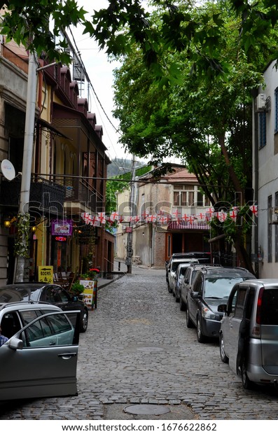 Tbilisi, Georgia - May 20, 2018: A quiet inner\
cobbled street with many parked cars and traditional architecture\
of cafes & hotels in central Tbilisi. Note the string of\
Georgian flags with red\
cross.