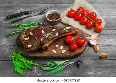 T-bone steak cow meat rib eye grilled on the board. Ingredients for T-bone steak cow meat rib eye grilled Tomatoes, rosemary, sage leaves, thyme, basil,  garlic, pepper. Gray wooden rustic background.