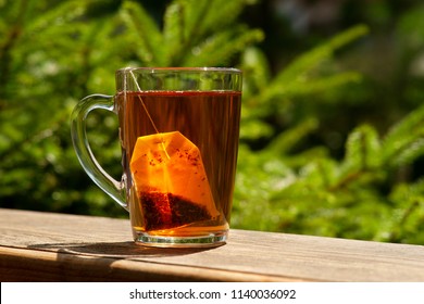 Tea And Tea Bag Under The Summer Sun. A Tea Bag In A Glass Mug On The Windowsill And Against The Background Of Green Summer Trees.