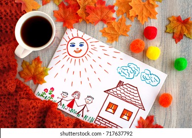 Tea And Drawing Of The Family. A Cup Of Tea And A Picture Of A Happy Joyful Family. A Tea Mug, Maple Leaves, Knitted Orange Shawl, Family Drawing Symbolize Coziness In The Autumn Season.