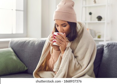 Tea to get better. Young woman wearing a knitted sweater and hat sitting at home on the sofa wrapped in a plaid drinking hot coffee or tea. Frozen woman will warm up in a cozy living room.