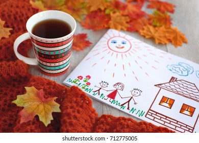 Tea And Happy Family. A Mug Of Tea And A Picture Of A Joyful And Friendly Family Create Coziness. Autumn Composition. Tea In Orange Colors. Fallen Leaves, Tea, Knitted Shawl And Family Drawing.