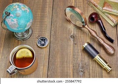 Tea Mag On Grunge Wood Table With Many Travel Objects. Compass, Spyglass, Vintage Notebooks, Bell, Retro Globe Map, Smoking Pipe, Magnifying Glass