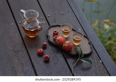 A tea set on a wooden table with glass pitcher, two glass cups, red fruits, possibly apples, and persimmons on a decorative tray, with green leaves. - Powered by Shutterstock