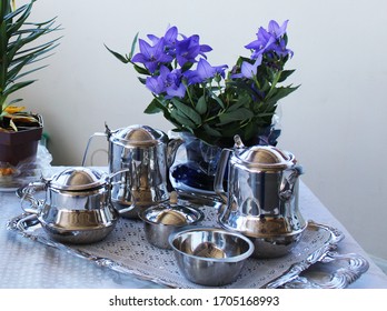 Tea set with silver objects on a silver tray. behind it is a vase of violet and purple flowers. everything is supported on table with white towel