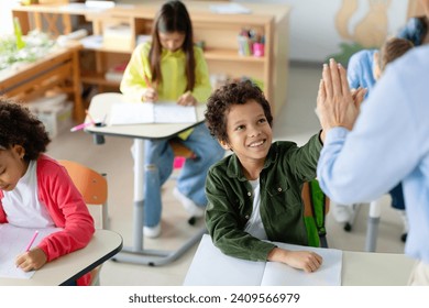 Teacher giving high five to schoolboy, achievement, success and mentorship for learning. Students with goals, knowledge and books with celebration at school desk - Powered by Shutterstock