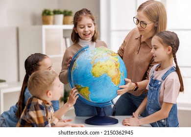 Teacher in glasses and positive children examining globe while standing around table in geography classroom during lesson at school - Powered by Shutterstock