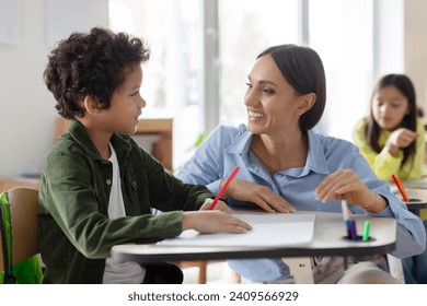 Teacher helping latin boy with assignment in elementary school, classroom support, scholarship and educator aid, explaining or helping male learner in class - Powered by Shutterstock