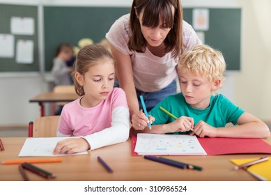 Teacher helping two small kids in the classroom as she leans over them to write something on the little boys work, both children watching with solemn faces
