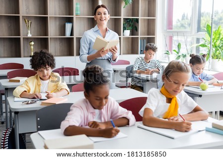 teacher holding book and looking at camera while multicultural pupils writing dictation in classroom