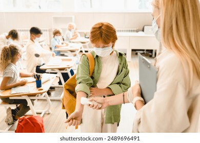 Teacher measuring temperature on schoolgirl`s wrist student classmate before lesson wearing protective medicine face masks against coronavirus Covid19 due to lockdown restrictions. - Powered by Shutterstock