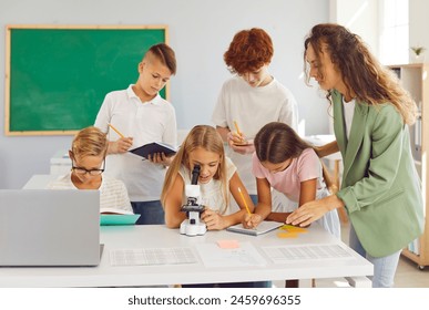 Teacher observes how girl student looks into microscope during biology lesson in primary school. Schoolchildren write down the information dictated by their classmate in their notebooks.  - Powered by Shutterstock