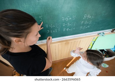 A teacher points to math problems on a chalkboard as a young student observes and participates. They are learning about calculations in a classroom with natural lighting.
