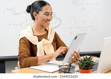 Teacher posing with tablet pc at elementary school. Young teacher of Latin appearance in cardigan, glasses holds a tablet in her hand. Looks at the device, prints and prepares for the lesson