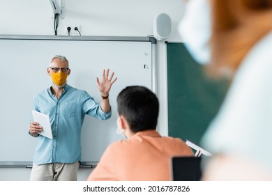 Teacher In Protective Mask Holding Digital Tablet And Showing Palm Of Hand Near Pupils