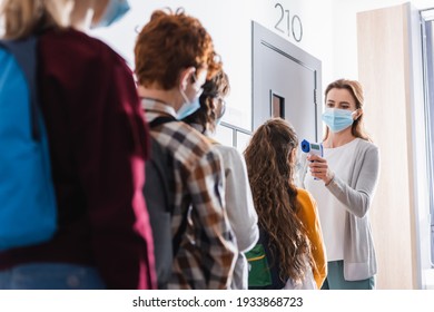 Teacher in protective mask measuring temperature of schoolchild near pupils on blurred foreground in hall - Powered by Shutterstock