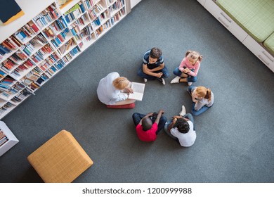 Profesor leyendo cuentos de hadas a los niños sentados en círculo en la biblioteca. Vista superior de los bibliotecarios sentados con cinco niños multiétnicos en el piso. Profesora leyendo libros a niñas y niños en la escuela.