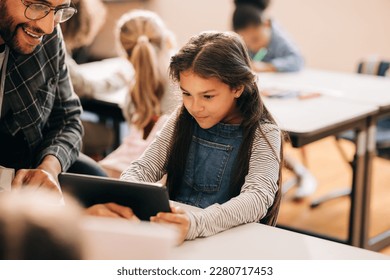 Teacher shows a student how to use a digital tablet in an elementary school. Male tutor teaches digital literacy and coding to young children. Child education and mentorship using modern technology. - Shutterstock ID 2280717453