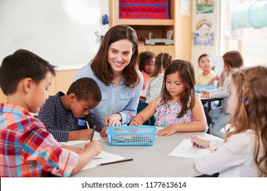 Teacher Sitting At Table With Young School Kids In Classroom