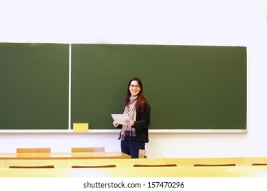 Teacher Smiling, Holding Paper In Empty Classroom, Blank Blackboard In Background