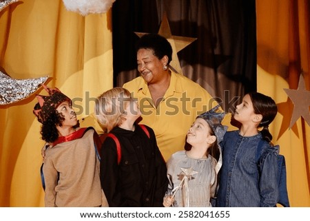 Teacher smiling warmly while interacting with group of children in classroom setting. Children looking up at teacher, appearing engaged and happy
