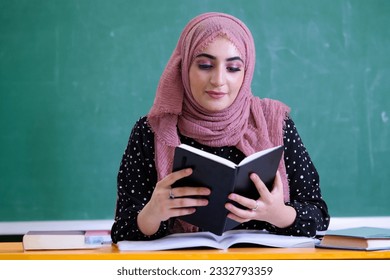 Teacher or student wearing Hijab scarf inside classroom with blackboard as background for education, university campus concept. Female holding a notebook at school