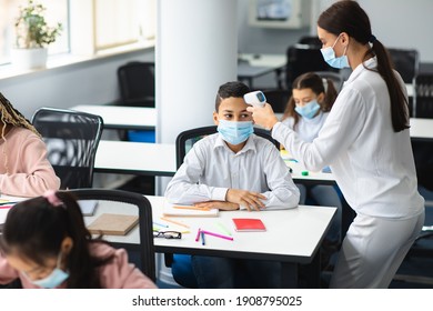 Teacher using digital medical contactless infrared electronic thermometer, measuring and checking body temperature, screening small boy in protective mask, preventing contagious diseases spread