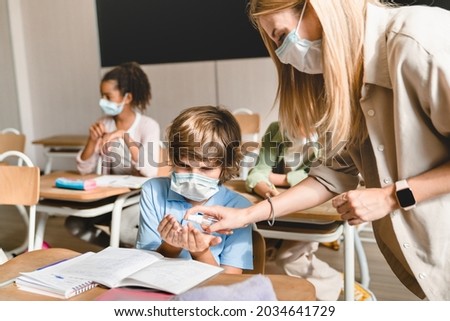 Teacher using sanitizer with her pupils schoolchildren kids students at lesson class in school wearing protective medicine masks against Covid19 coronavirus. School during pandemic