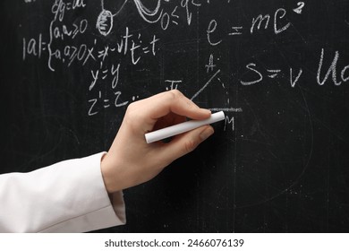 Teacher writing physical formulas with chalk on black chalkboard, closeup
