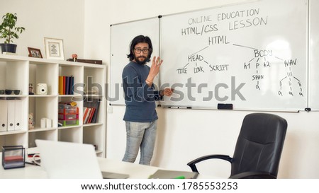 Teaching code programming. Young bearded male teacher pointing at whiteboard and explaining how to code HTML CSS, giving lesson online at home. Focus on man. E-learning. Distance education. Stay home
